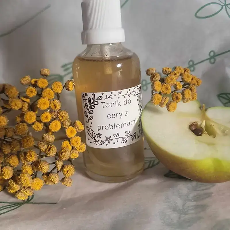 buteleczka toniku do twarzy na jasnym tle w towarzystwie żółtych kwiatów oraz przekrojonego jabłka