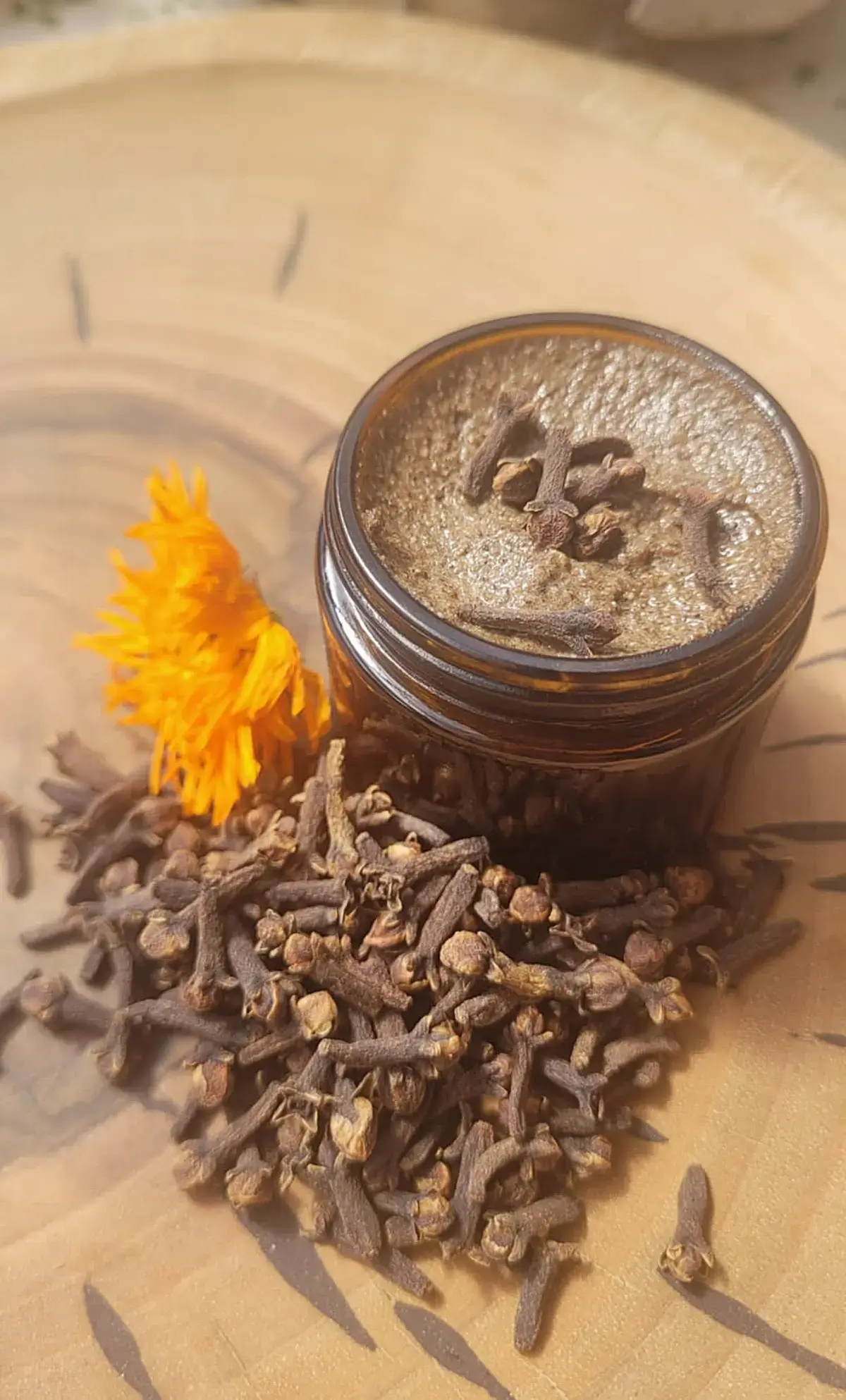 słoiczek goździkowego peelingu do twarzy na drewnianym tle w otoczeniu goździków i suszonych kwiatów