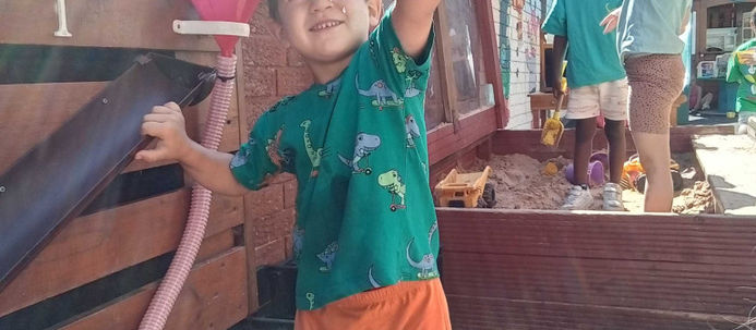 A young child wearing a green T-shirt is joyously holding up a flower shaped toy, smiling. He is standing next to a water feature, with a red water tray in front of him. There are a couple other children standing in the background in a sandpit. Playful, nursery environment.