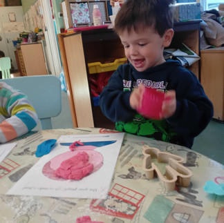 a brown haired little boy is happily playing with red play dough, there is a face templeta on the table in fron of him with red play dough on it