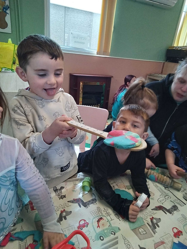 A happy little boy holding a wooden spoon and balancing a batch of colourful playdough on it. Other children ext to him looking at him.