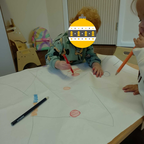 a baby holding a pencil is making mark on a large piece of paper that has a Christmas tree drawn on it