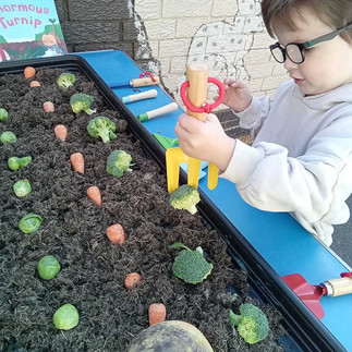 a little boy is holding a yellow rake over a row of vegetables dug into a tray full of soil