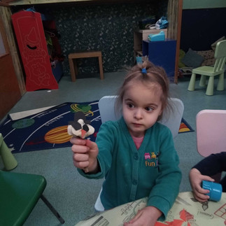 a little girl wearing Fun Box Day Nursery uniform is holding up a playdough snowmen she has just made, there is wooden play house and some chairs in the background