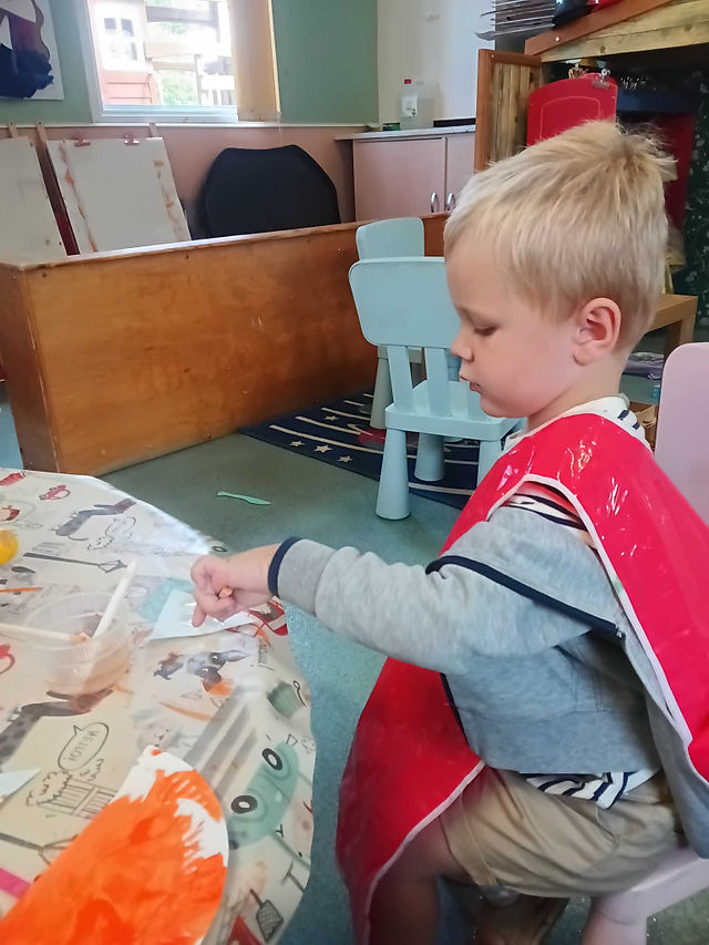 a blonde haired little boy, wearing a red tabard is painting a piece of card with orange paint, there are blue chairs and easels in the background