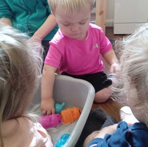 a group of toddlers reaching into a gray bowl that has bubbly water and bath toys in it, a little boy wearing pink t-shirt is in the middle