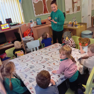 a group of children sitting around a table with white table cloth on it, a teacher wearing Fun Box Day Nursery uniform is holding up a small jar of marmalade, showing to them