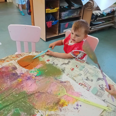 a little boy is using a fly swatter to splatter paint on a piece of paper, a unit with different coloured drawers is behind him, nursery environment