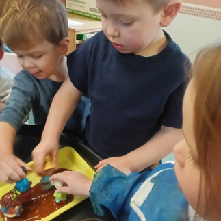 three young children are putting small toy people into a yellow tray filled with brown paint
