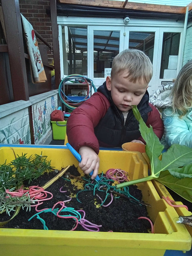 blonde haired little boy in a red jacket is using tweezers to explore coloured pasta in a sensory train, outdoor setting at nursery in Totton