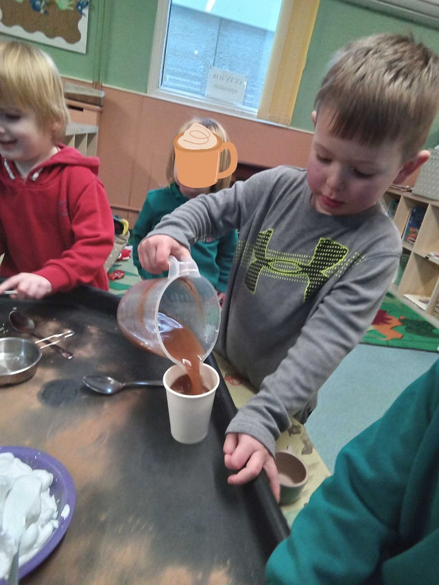 a little boy is using a see-through plastic jug to pour hot chocolate into a paper cup, there are other children enganged in the same activity around the builders tray