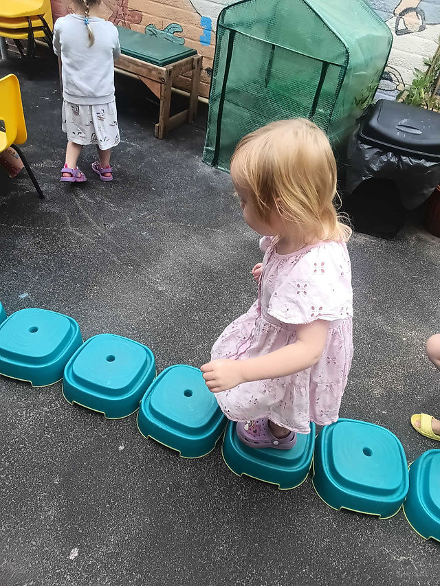 a young little girl wearing a summer dress is balancing carefully on green, plastic stepping stones in a nursery garden