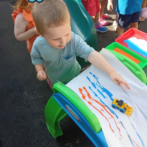 a little boy is letting go of a small yellow truck, that was dipped in paint, the truck is running down on a small slide covered with paper