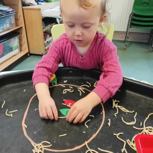 little girl wearing a pink jumper is makin a face in a sensory tray using spaghetti and colourful paper shapes