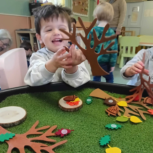 Smiling child proudly showing a felt tree at the autumn sensory table surrounded by colourful leaves and acorns during Fun Boxās āAll the Leaves Are Falling Downā activity.