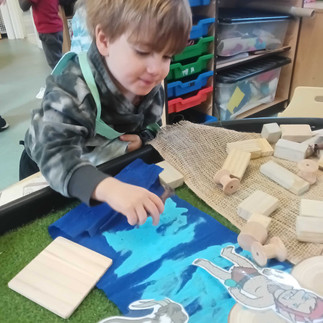 a little boy is playing with wooden loose parts in a sensory tray, there is a Troll and and Billy Goat cutout picture on the tray