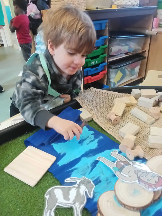 a little boy is playing with wooden loose parts in a sensory tray, there is a Troll and and Billy Goat cutout picture on the tray