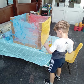 a young boy is using a paintbrush to decorate a large cube on a blue table, nursery playground behind him