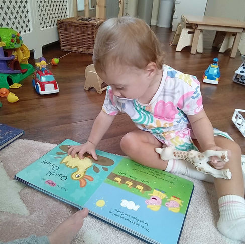 a baby girl is looking at a Peppa Pig storybook, touching the fluffy duck picture, there are toys , a large treasure box, a low wooden baby table in the background, nursery environment
