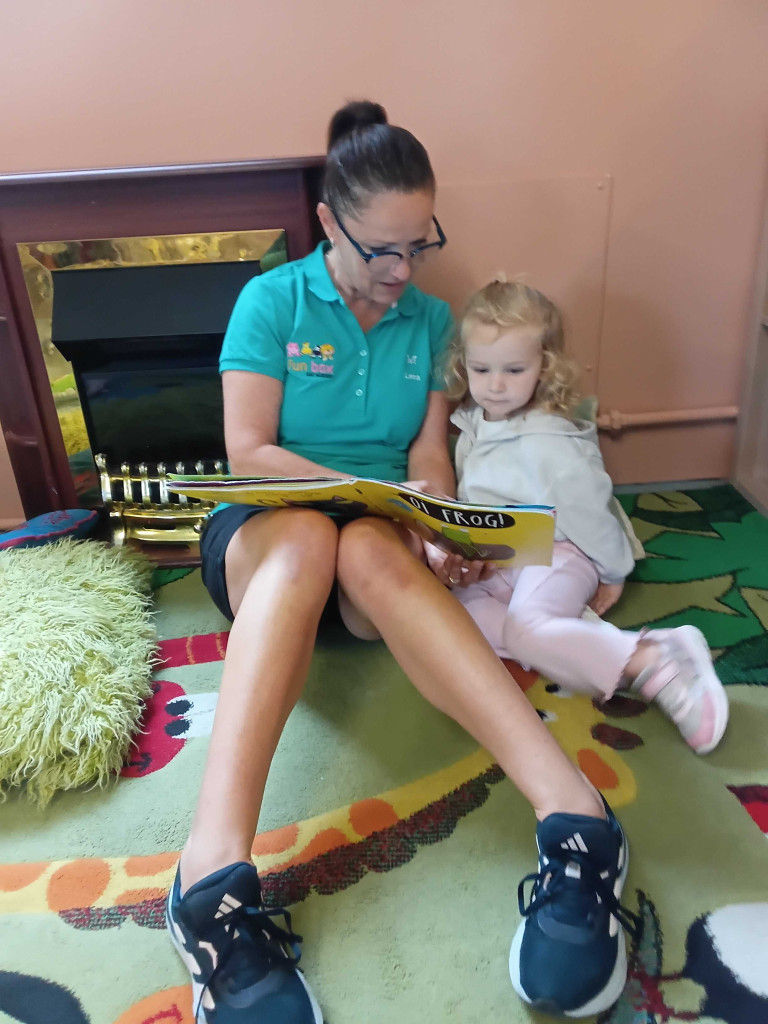 A Fun Box Day Nursery teacher and young girl sit on a colorful rug reading a Frog! book by a fireplace, sharing a calm storytime.