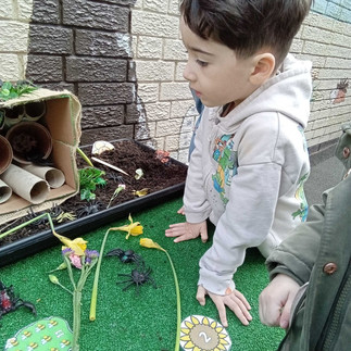 a black haired little boy is looking at a bug hotel set up a fake grass table, several toy insects in front of the hotel
