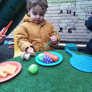 a little boy is using plastic cutlery to pretend to cut up toy fruits and vegetables in a nursery garden