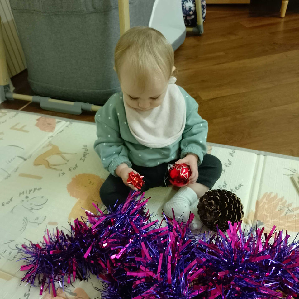 a little baby is exploring a bauble, tinsel, bow and pine cone on a beige carpet