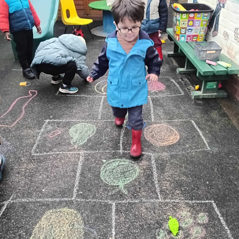 a little boy wearing glasses and blue coat is walking on a chalk drawn game of fruit hoptscotch