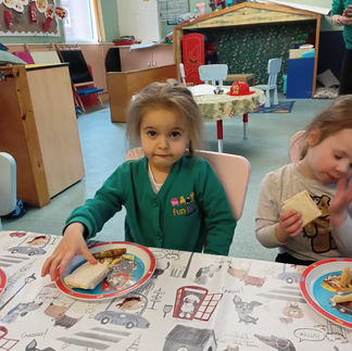 a little girl wearing Fun Box Day Nursery uniform is looking at the camera as she is reaching for a sandwich