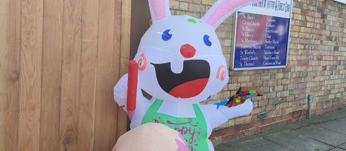 Inflatable Easter bunny with a green apron and oversized pink egg stands by a brick building and wooden gate. Blue and white sign in background.