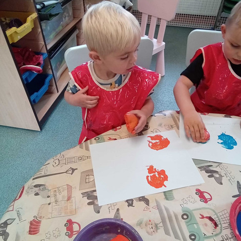 blonde haired little boy wearing a red apron is holding an orange he is using to print on paper using different coloured paint