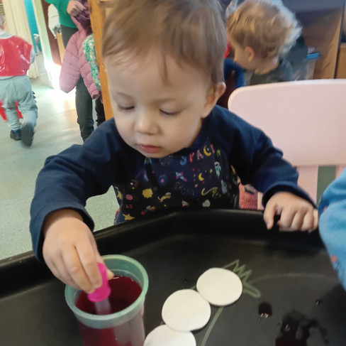 a little toddler is dipping a pipette into a tub with red water in it, there are a few round cotton pads in front of him on a black sensory tray, other children int the background