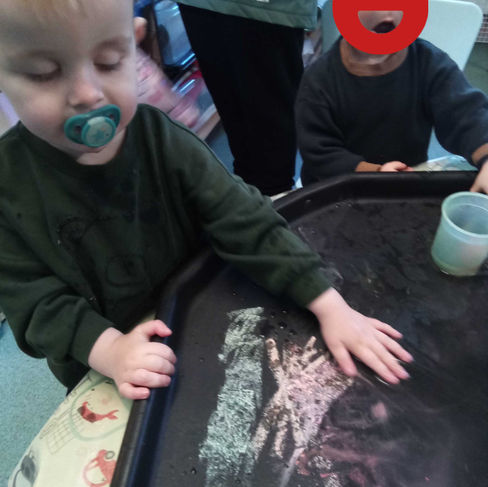 a young child with a dummy in his mouth is reaching onto a sensory tray that has chalk marks on it