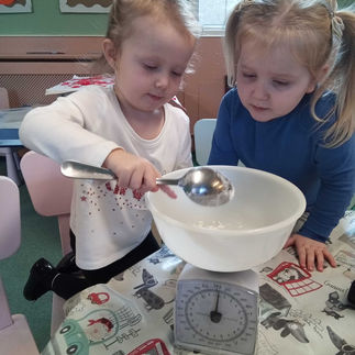 two little girls are scooping flour with a metal spoon into a plastic bowl