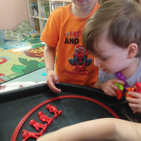 two young children are looking at a bunch of red figurines carefully laid out in a red circle on the table