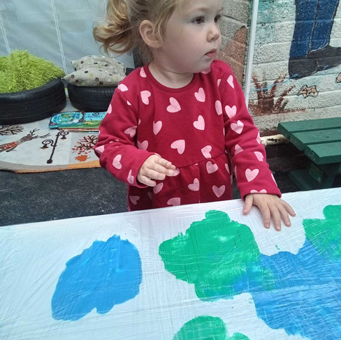 a toddler is looking on as there is a large piece of paper in front of them with green and blue paint under clingfilm
