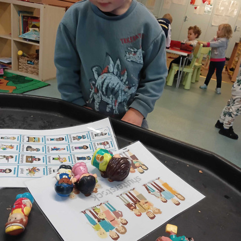 A young child stands at a play table observing small world people figures placed on top of a matching picture sheet. The activity supports learning about families, professions and diversity through hands-on exploration and imaginative play.