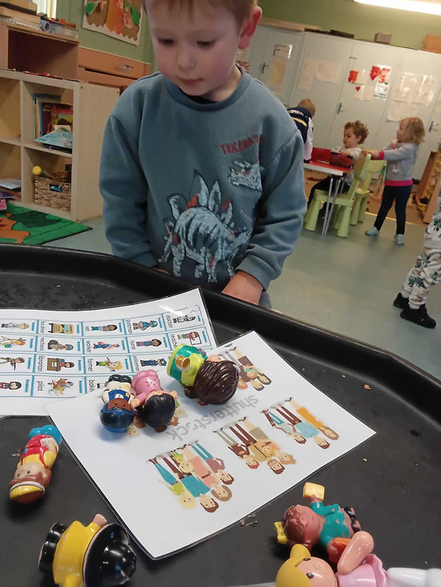A young child stands at a play table observing small world people figures placed on top of a matching picture sheet. The activity supports learning about families, professions and diversity through hands-on exploration and imaginative play.