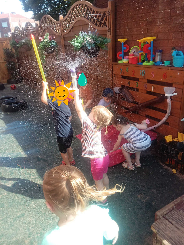 a group of children are blasting water into the air from giant water syringes at Fun Box Day Nursery's garden
