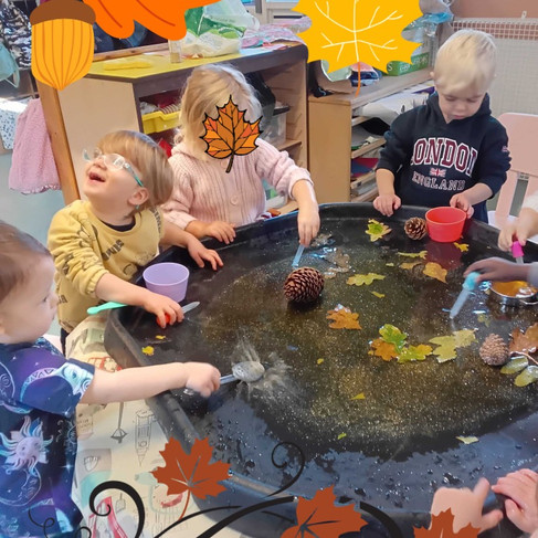 Group of children enjoying autumn sensory water play with leaves, pinecones and glitter, smiling and splashing together at Fun Box Day Nurseryās autumn activity.