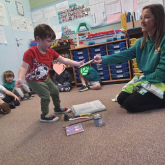 A young child stands in front of a teacher during a memory game with household objects placed on the floor. The child is pointing and thinking carefully, trying to identify which item has been removed from the original lineup, practising observation and recall skills through play.
