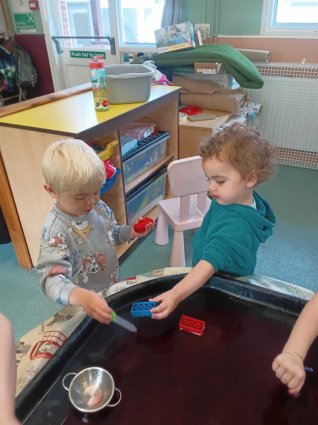 two young children are using pipettes in a black sensory tray filled with red water, in the background there is a large shelving unit and a door
