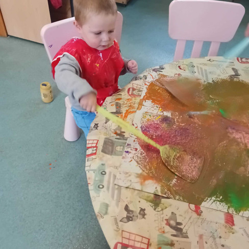 a young child wearing a tabard is splatting paint on paper in Fun Box Day Nursery