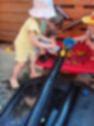 Children in sun hats play with water and toys at a red-and-black outdoor water table by a wooden fence.