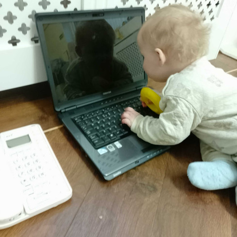 a baby boy is looking at his reflection at the monitor of an old laptop on the flor in front of a white cabinet, there is an old fashioned telephone next to the laptop