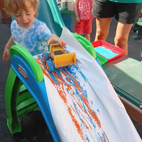 a toddler wearing a light blue t-shirt is holding a toy excavator, the toy is dipped in paint, the child is letting the card down on a small slide covered with paper, other children in the background