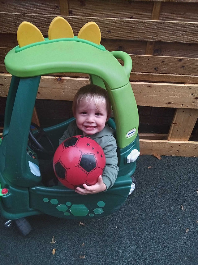 Smiling child sitting inside an outdoor play car while holding a red football at Fun Box nursery.