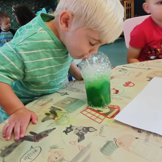 a little boy is blowing bubbles into a pot containing a green liquid, colourful table cloth on the table