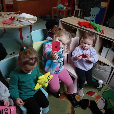 a group of young children sitting on small charies ar holding paper poppies in their hands, one of them is holding up their poppy to the camera