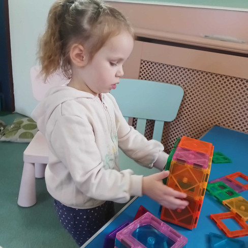 a little girls is focusing very hard as she is buillding a tower from magnetiles on a blue table with additional magnetiles around her